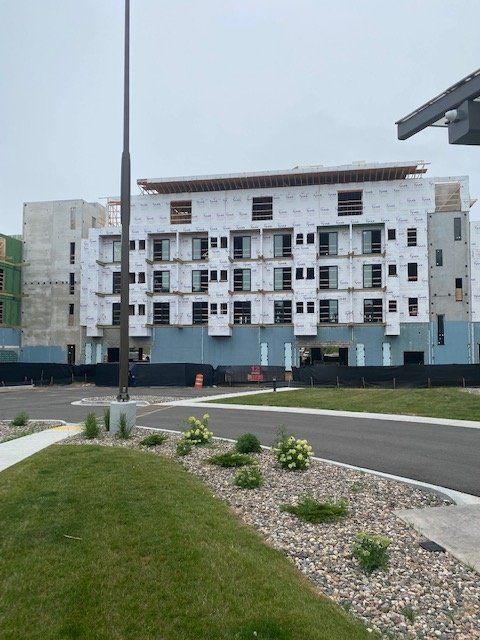 Building under construction, gray siding, many window openings, blue base, black fence, overcast sky.