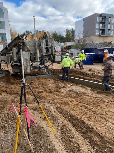 Construction workers pouring concrete curb at a building site. A concrete mixer and survey equipment are present.