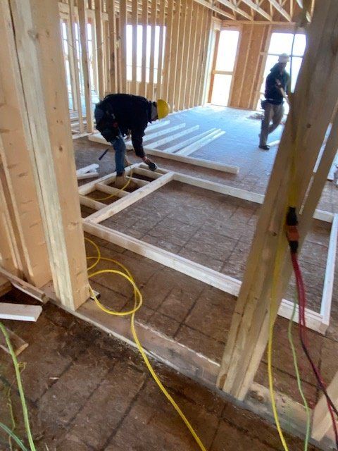 Construction workers building a wooden frame on a plywood floor. Yellow and red power cords visible.