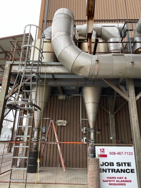 Industrial site exterior with metal piping, two conical hoppers, and a ladder. Job site entrance sign.