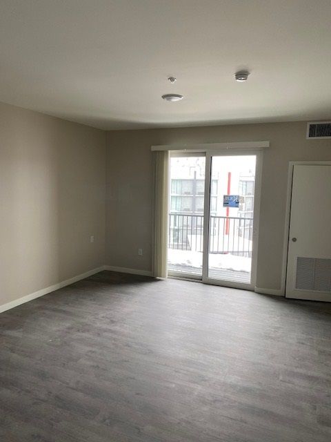 Empty room with sliding glass door and door to the right. Beige walls, gray flooring.