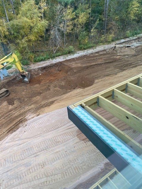Construction site with wooden deck framing in foreground. Dirt and an excavator are visible in the background.