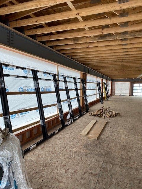 Interior of a building under construction, featuring large black-framed windows, wood beams, and exposed plywood flooring.