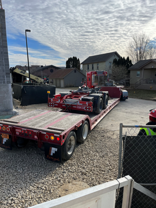 Red flatbed truck with oversized load, parked outdoors under cloudy sky.