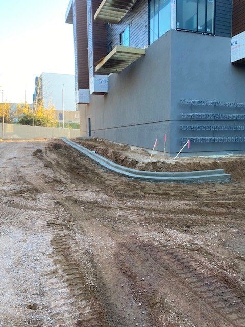 Construction site with a concrete curb in front of a building. Dirt and tire tracks visible.