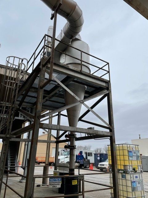 Metal industrial cyclone separator on a platform, outside. Steel frame structure, pipe, and blue barrels.