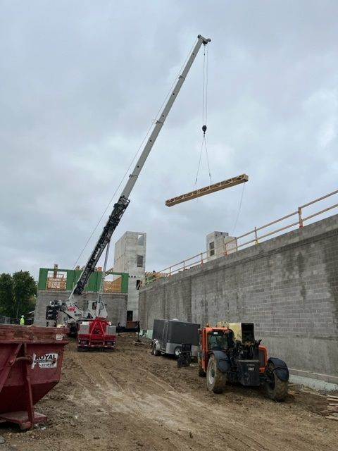 Construction site with crane lifting a wooden beam. A building is under construction, cloudy sky.