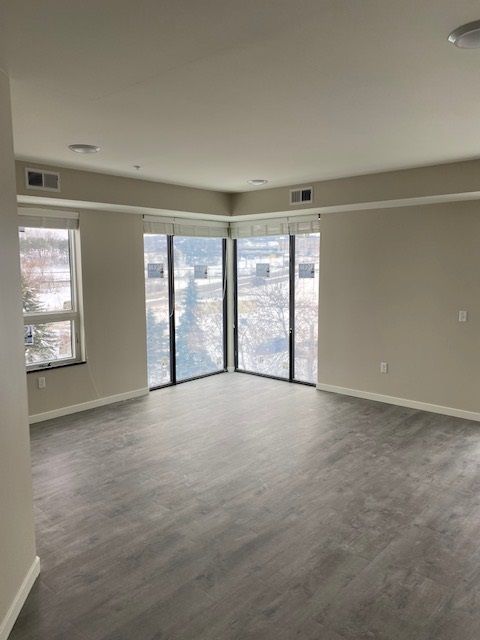 Empty room with light gray flooring and neutral walls. Large windows with shades offer a view of snow-covered trees.