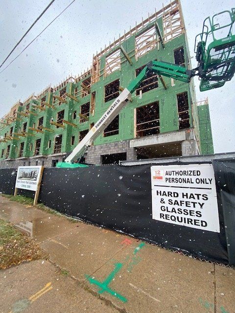 Construction site: multi-story building frame with green siding, green lift, black fence, and safety sign. Snow is falling.