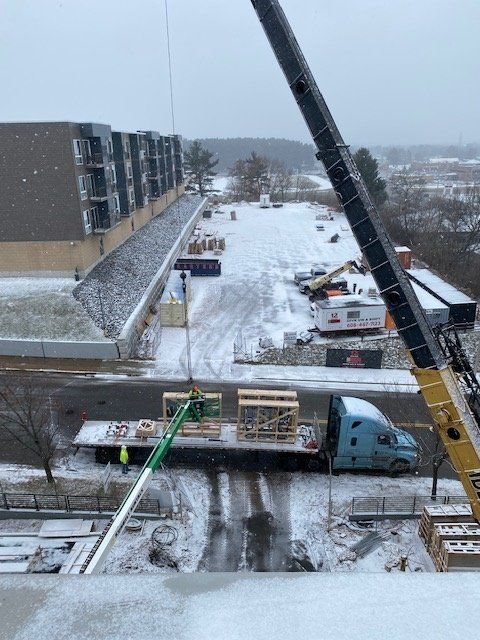 Crane lifting wooden crates from a flatbed truck at a construction site covered in snow.
