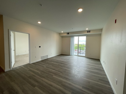 Empty living room with gray wood-look flooring, white walls, door, and a sliding glass door to a balcony.