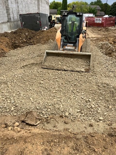 Skid steer loader smoothing gravel on a construction site; dirt and buildings in background.