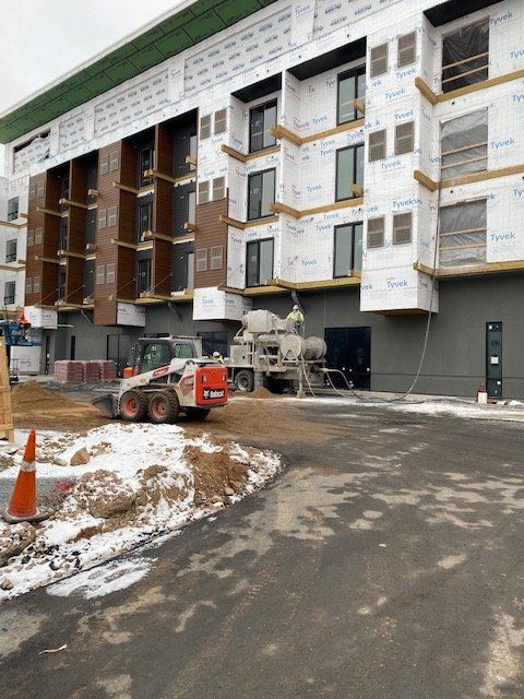 Construction site with a partially-built building, a skid steer, and a concrete mixer on a paved surface.