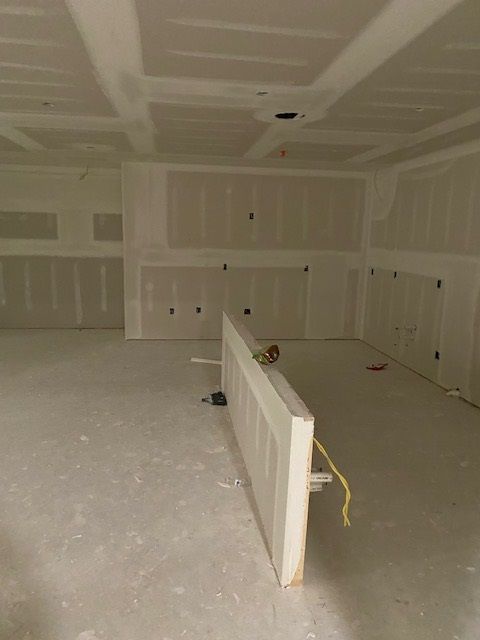 Interior room under construction, with drywall walls and ceiling. Door leaning against a wall, unfinished flooring.