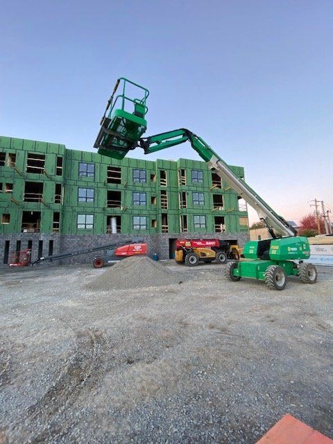 Green construction lift extended, working on a wood-framed building under construction. Gravel and equipment on site.