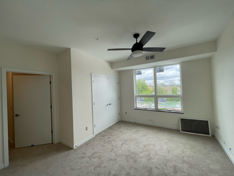 Empty bedroom with light beige walls, carpet, ceiling fan, and large window.