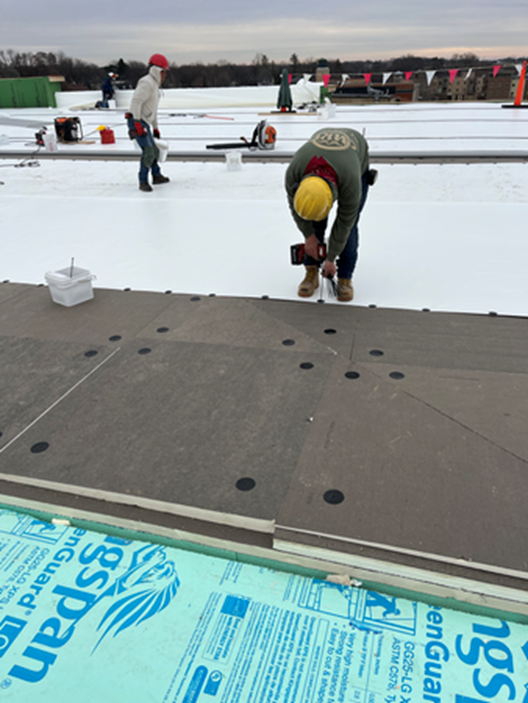Construction workers on a roof, installing insulation. One drills, others prepare materials. Cloudy sky in the background.