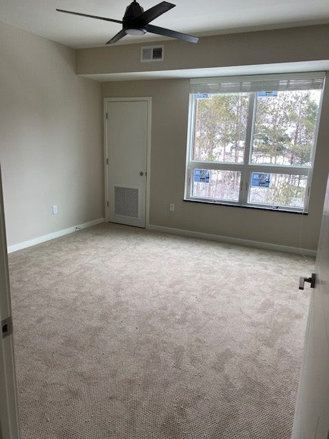Empty room with beige walls and carpet, a window, door, and ceiling fan.