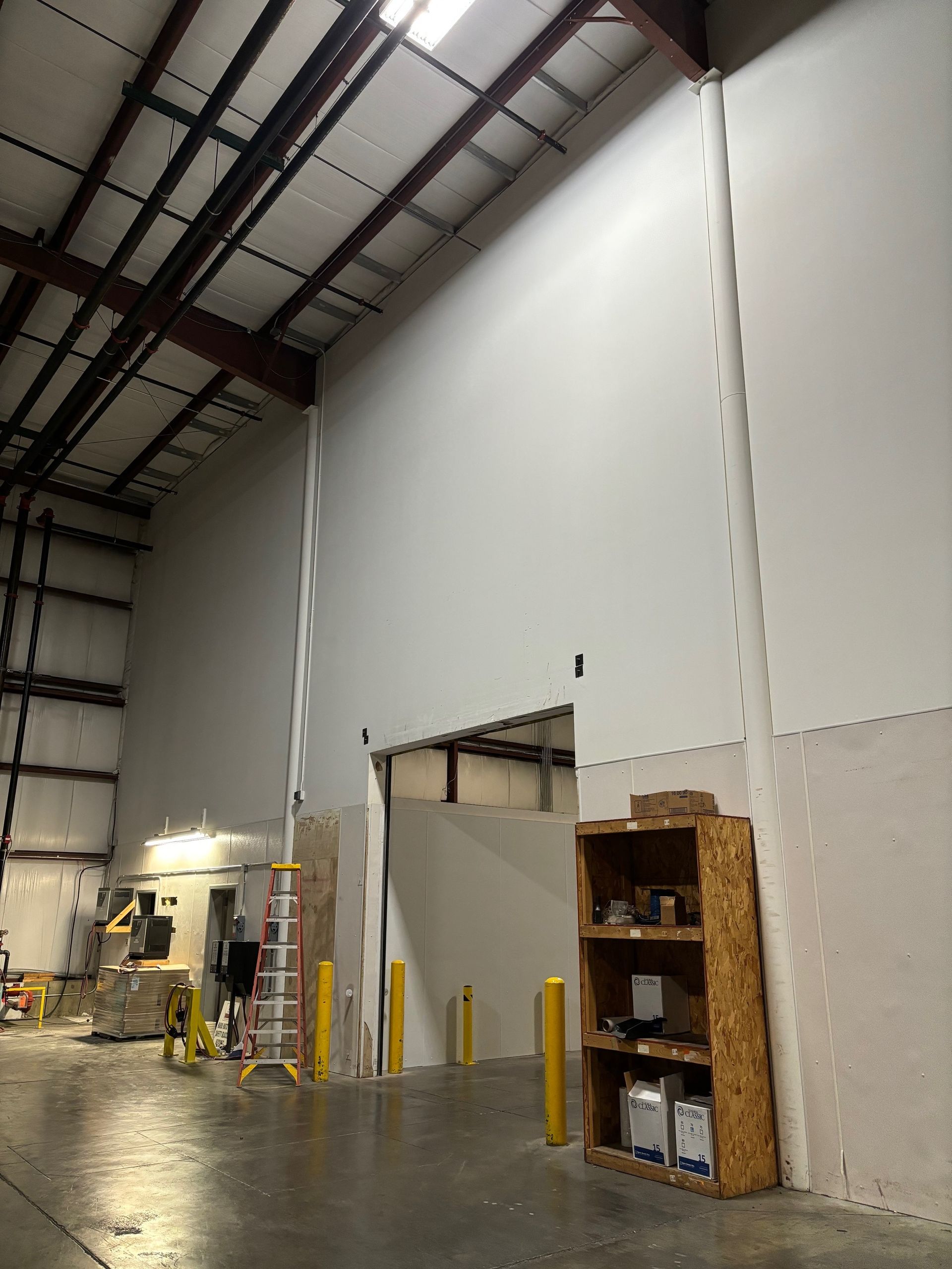 Warehouse interior with tall white wall, open doorway, shelving unit, and ladder.