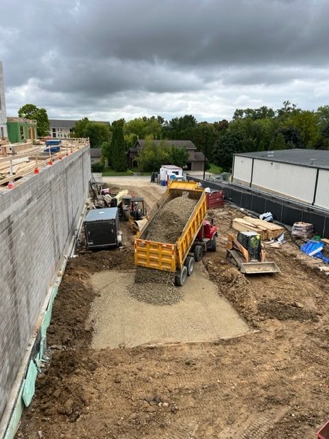 Construction site with dump truck unloading gravel. Backhoe, other equipment, and new building under construction are visible.