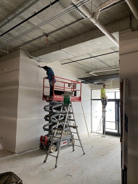Construction workers on lifts and stilts drywalling a building interior.