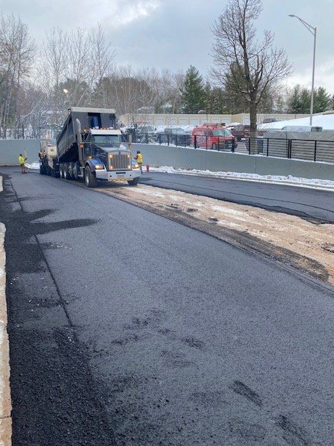 A dump truck unloading asphalt on a road construction site. Workers in safety vests are present.