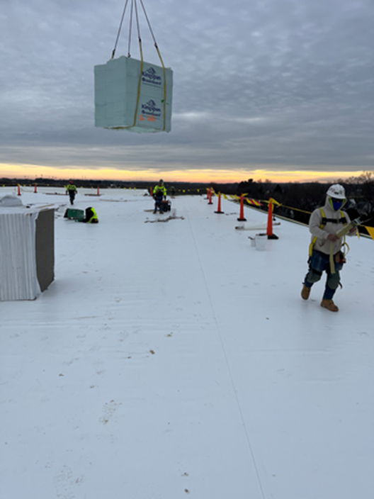 Workers installing a roofing material on a flat roof. A large bundle is being lifted by a crane.