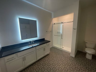 Bathroom with white vanity, black countertop, lit mirror, glass shower, and patterned floor.