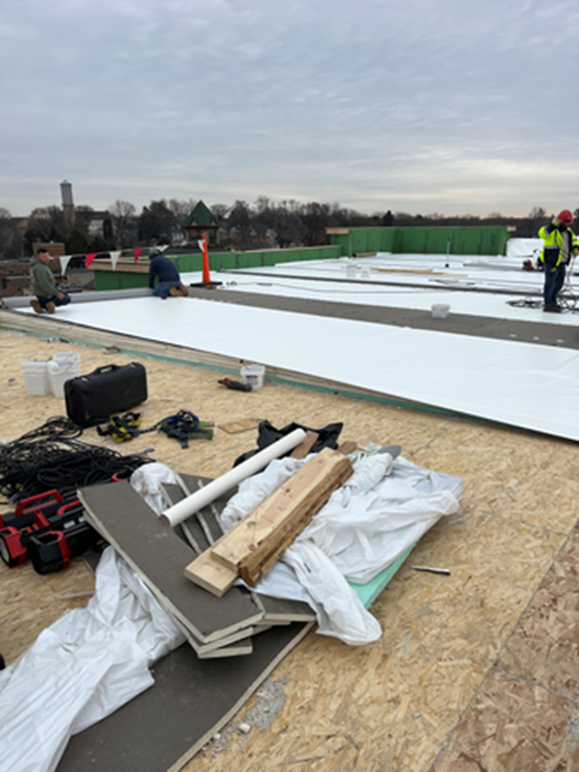 Construction workers on a rooftop installing white roofing material, cloudy sky.
