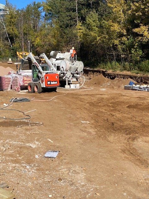 Construction site with Bobcat, concrete mixer, and materials. Worker on mixer, dirt ground, trees in background.