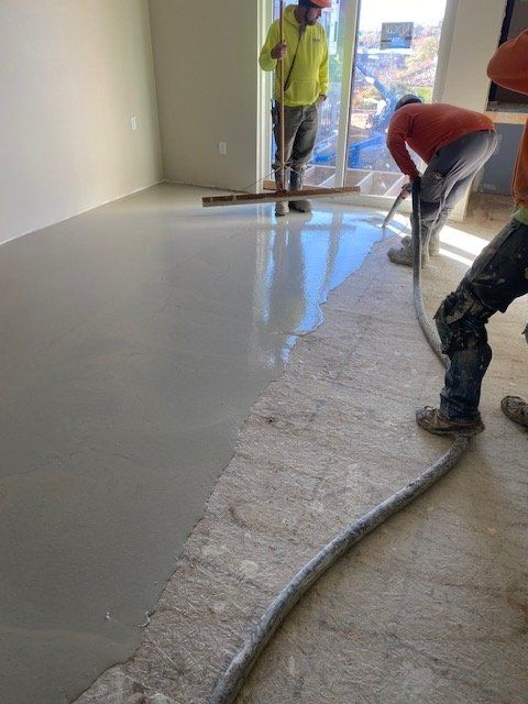 Construction workers applying a smooth, gray concrete floor. One uses a pump, another levels with a bar. Indoor setting.