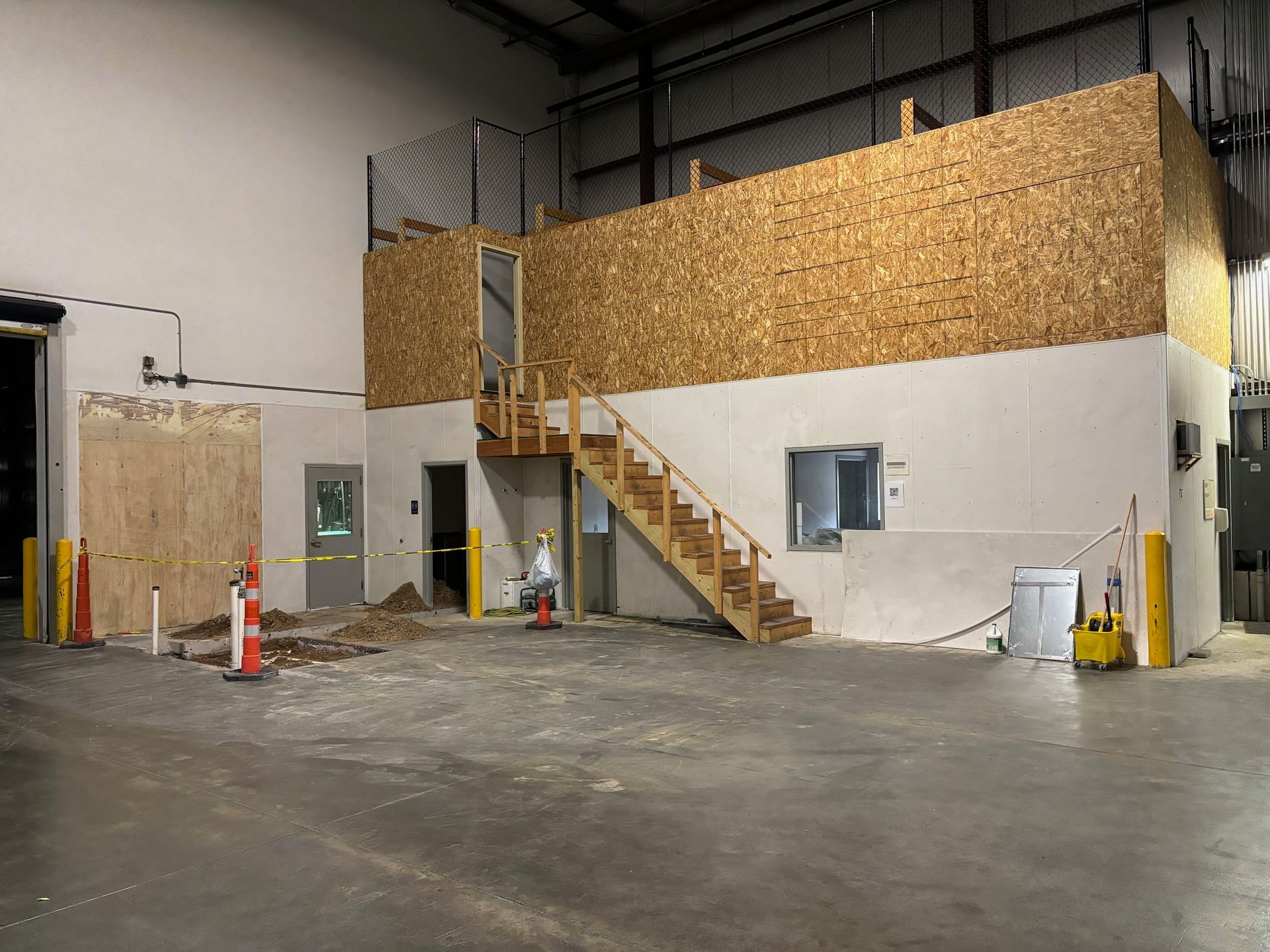 Interior view of a warehouse with a two-story wooden structure under construction, a staircase, and open doorway.