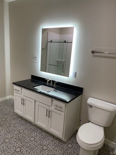 Bathroom with white vanity, black countertop, lit mirror, toilet, and patterned floor.