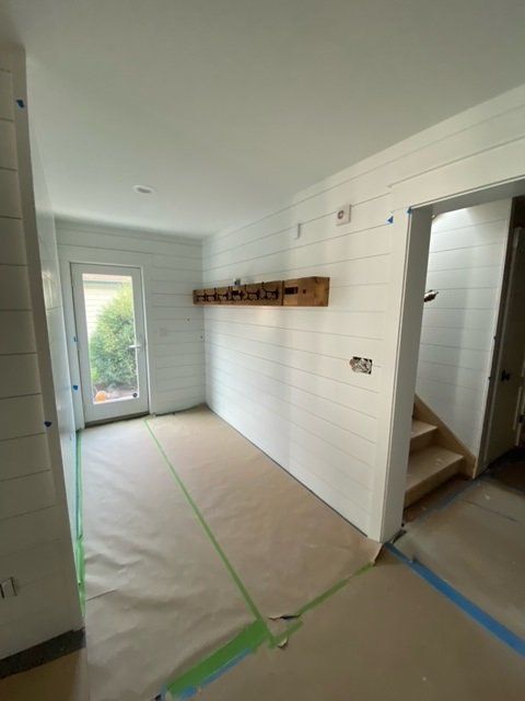 White-walled room under construction with wood shelf. Door to outside, stairs to upper level, and paper floor covering.