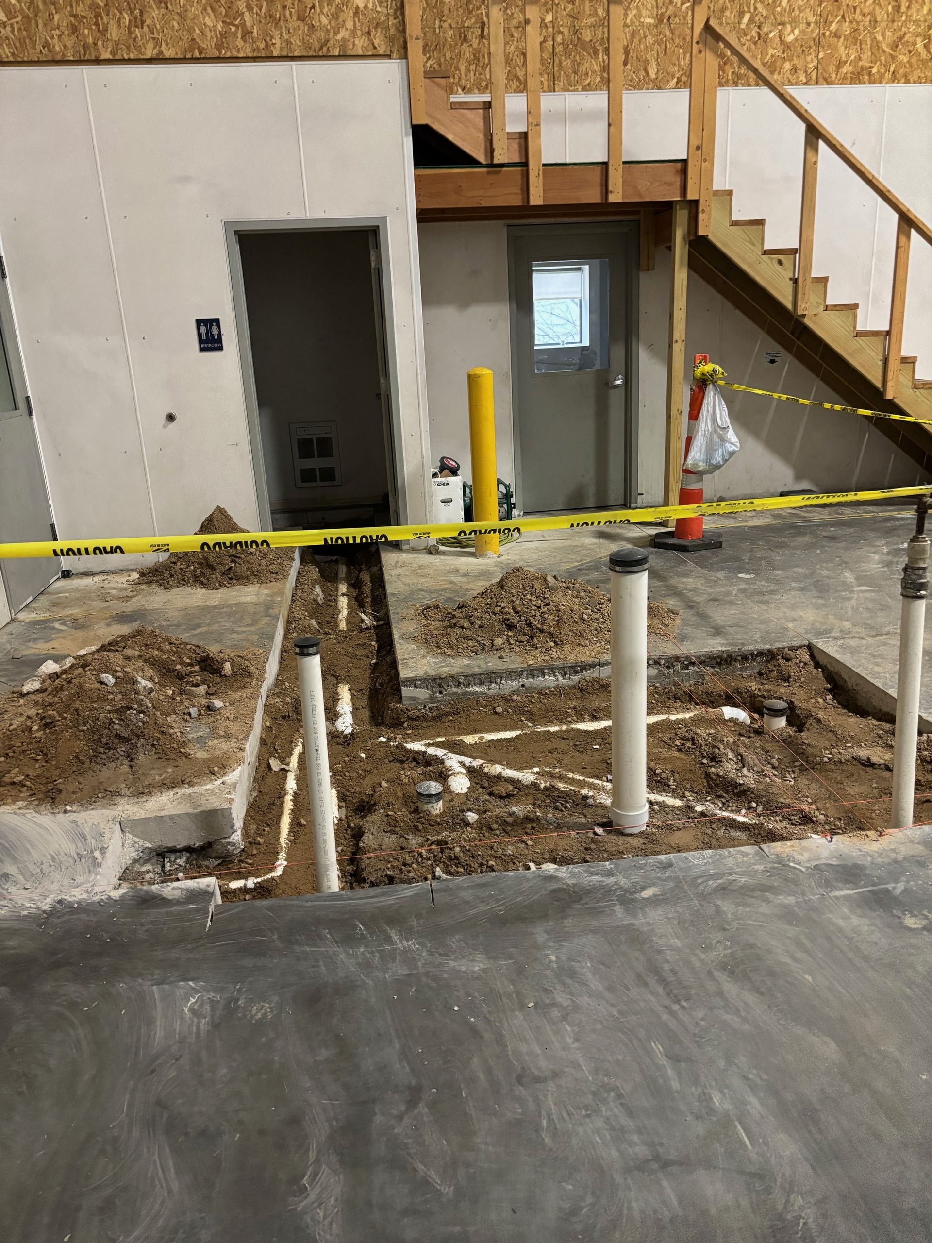 Construction site: Exposed pipes in dirt trench, caution tape, doorway, stairs.