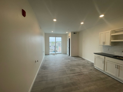Empty apartment room with white walls, carpet, and a small kitchen area.