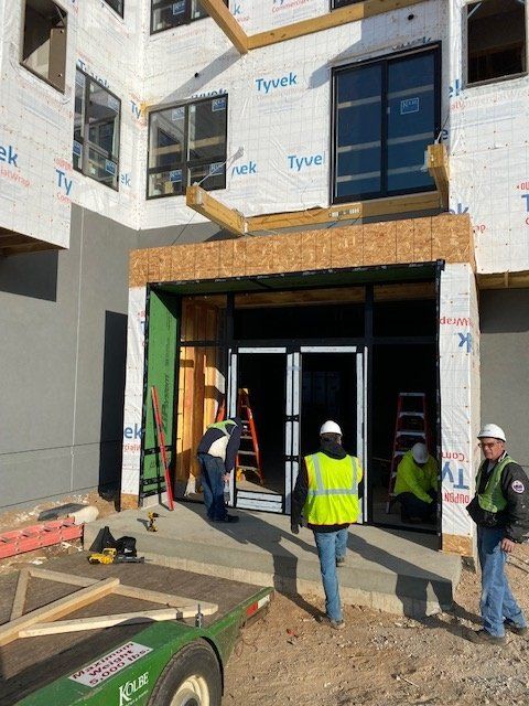 Construction workers at building entrance; scaffolding, safety vests, open doorway, tools, Tyvek siding.
