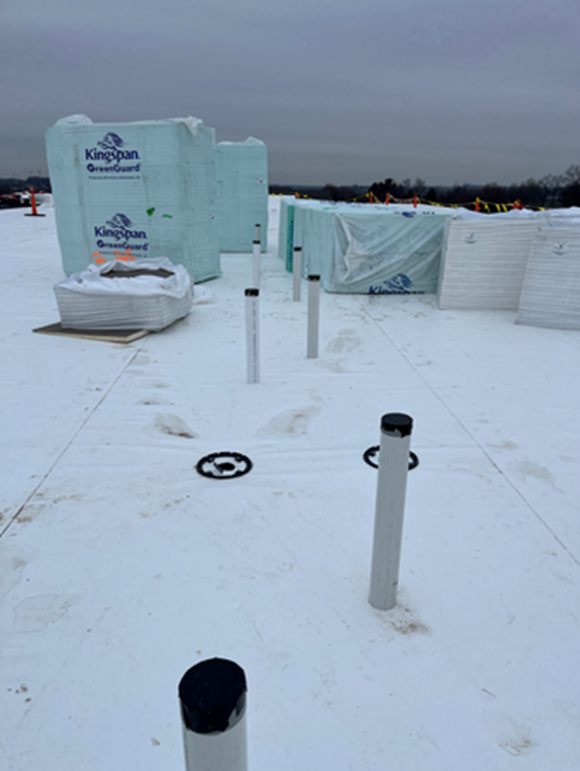 Flat white roof with building materials. Several white poles and a drain are visible. Overcast sky.