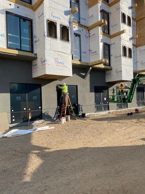 Construction site: workers install a window on a building's gray exterior, using a ladder and lift. Dry dirt ground in front.