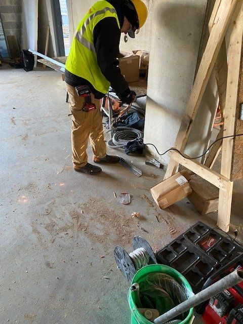 Construction worker cutting metal pipe indoors; concrete floor; yellow vest; safety helmet.