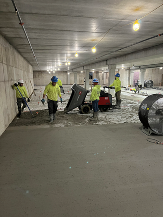 Construction workers pouring concrete in a large, unfinished building. They are wearing hard hats and working on the floor.