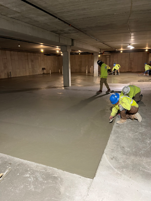Construction workers smoothing wet concrete floor in a large, unfinished concrete room.
