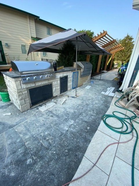 Outdoor kitchen with grill, fridge, and overhead shade, under construction.