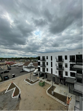 Modern apartment building with balconies under a cloudy sky. Driveway, green dumpster, and nearby businesses.