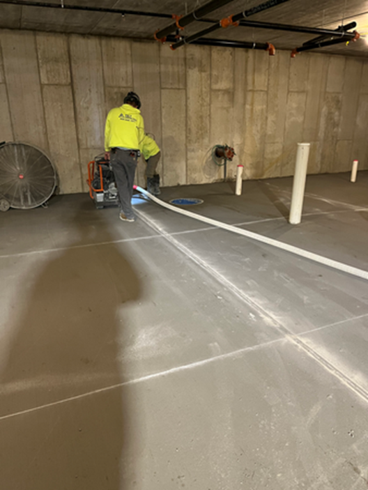Two workers in a concrete room pumping something into a long white hose laid across the floor.