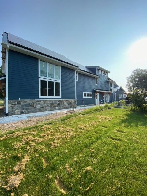 Blue-sided house with stone base and large windows, solar panels on the roof, set in a grassy yard under a bright sky.