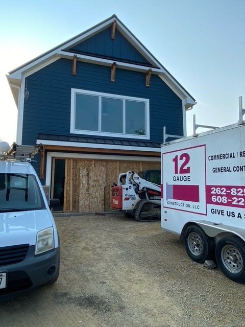 House under construction with a white van, construction equipment, and a company trailer. Blue siding, exposed wood framing.