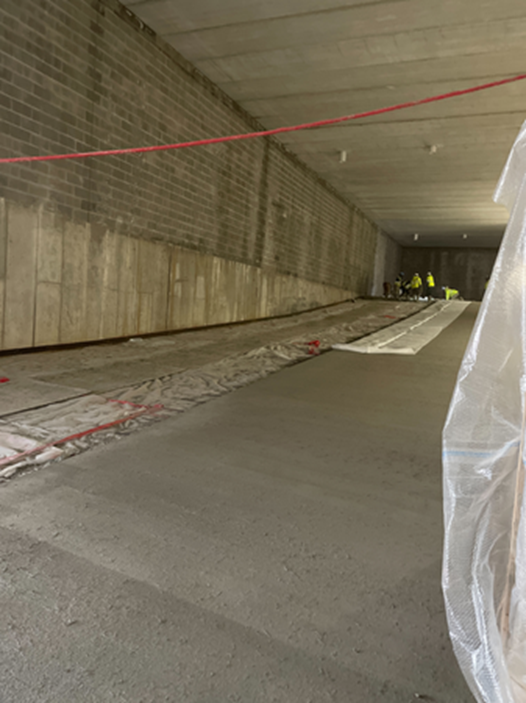 Construction work in tunnel; concrete floor, brick wall. Two workers in vests visible in the distance.