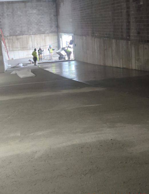 Workers smoothing concrete floor in a large, unfinished building. Sunlight streams in from a doorway.