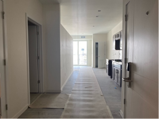 Hallway in a modern apartment, partially constructed. White walls, flooring covered in paper, kitchen in the distance.
